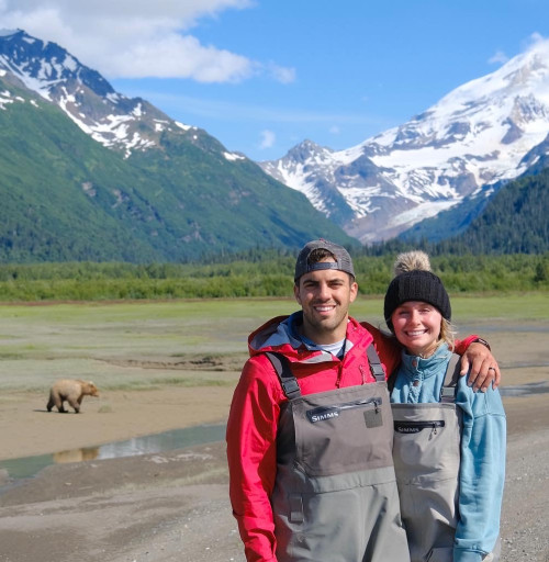 Tessa Wisniewski鈥�17, 鈥�19 and her husband standing on a shoreline with a bear and mountains behind them